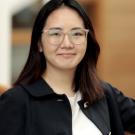 Portrait photograph of young woman with glasses, black jacket, blurred wooden background